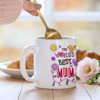 White mug with 'The World's Best Mum' text and colorful graphics, being stirred with a gold spoon.