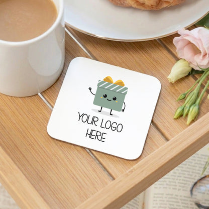 A square white coaster with rounded corners sits on a wooden table, printed with a cute gift box character and the text 'YOUR LOGO HERE'. A white mug, a plate with a pastry, and pink flowers are arranged around it in a styled setting.