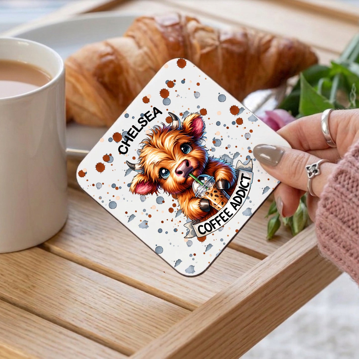 Card with a dog illustration and text 'Coffee Addict' held by a person over a cup of coffee and croissant.