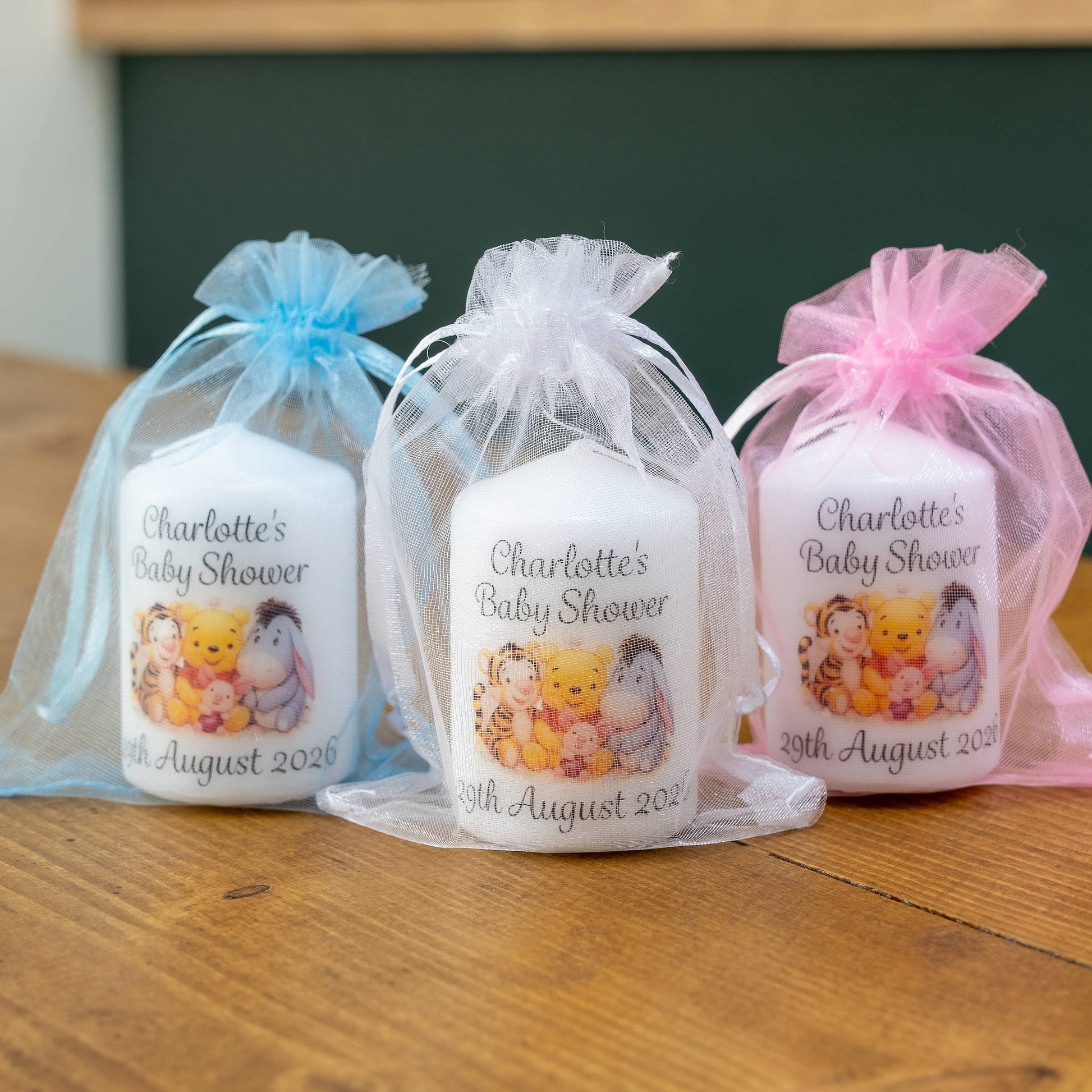 Three personalised baby shower candles in white wax packets, each tied with a sheer organza bag in light blue, white, and pink. The candles feature a printed illustration of Winnie the Pooh and other friends, with the text 'Charlotte's Baby Shower' and the date '20th August 2021'.