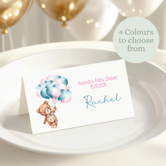 Baby shower place card with teddy bear and balloons on a white plate with gold balloons in the background.