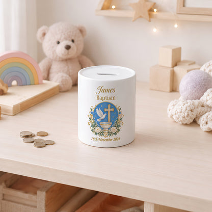 A white ceramic baptism money box with a gold slot on top, personalised with the name 'James Baptism' and a blue dove emblem, sitting on a light wooden table in a child's room with toys in the background.