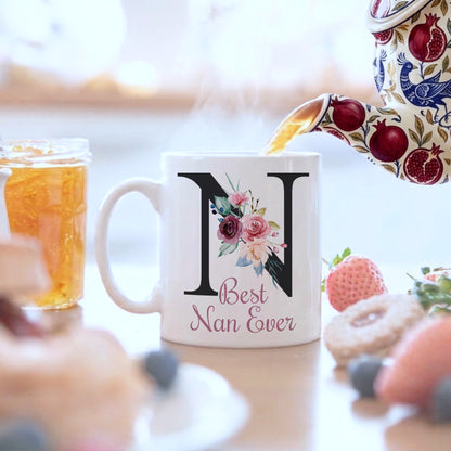 A white ceramic mug with a large black letter 'N' and floral design, printed with 'Best Nan Ever' in pink script. A patterned teapot pours tea into the mug, with a blurred breakfast table and food items in the background.