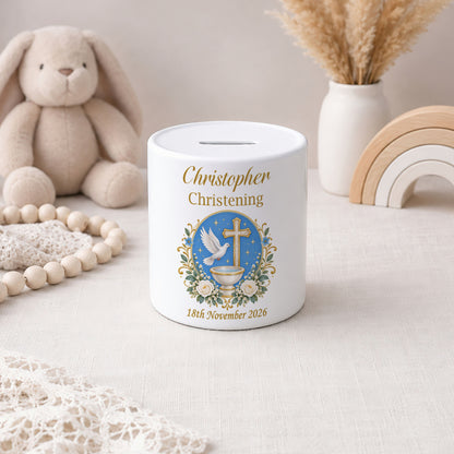 A white ceramic baby christening money box with a printed blue, gold, and white illustration of a dove, cross, and flowers, personalized with the name 'Christopher' and date '10th November 2026'. The box is styled on a linen surface with a plush bunny, wooden beads, and a small vase in the background.