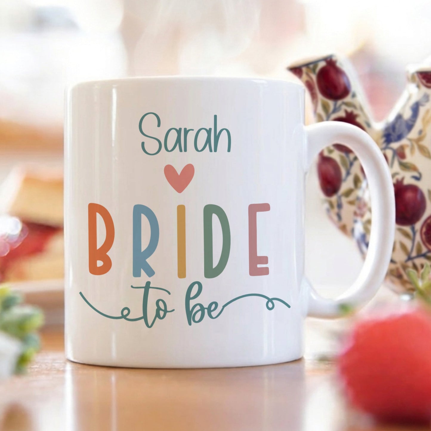 White mug with 'Sarah Bride to be' text on a blurred background of tea set and fruits