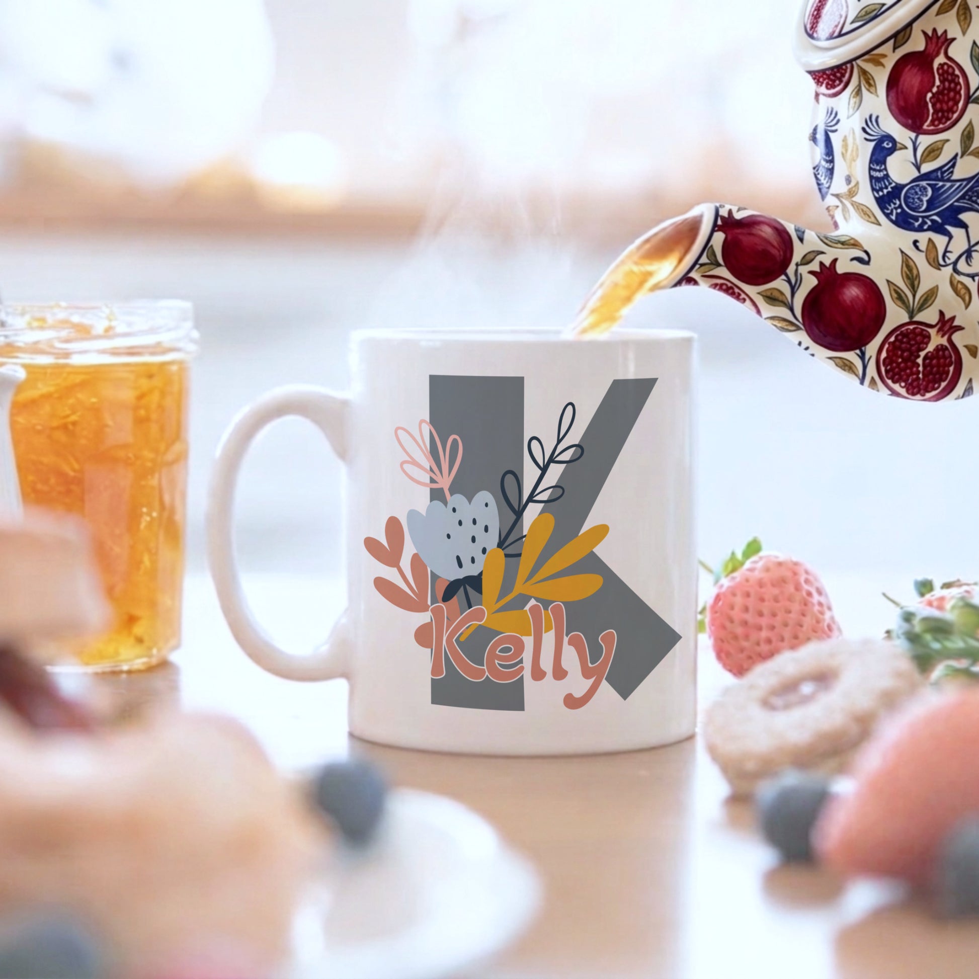 Mug with 'Kelly' design being filled with tea, surrounded by food items on a blurred background