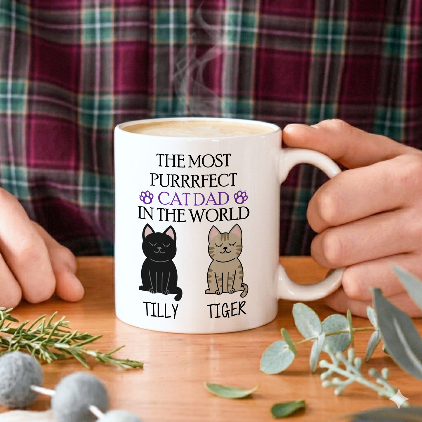 A white ceramic mug with a humorous cat dad graphic, held by hands against a plaid shirt background on a wooden table with decorative greenery.