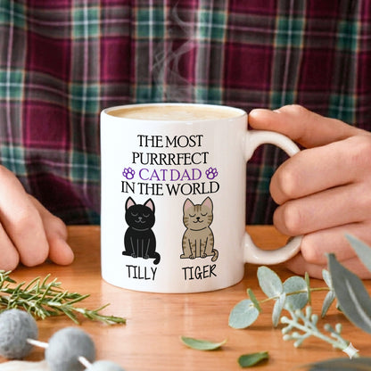 A white ceramic mug with a humorous cat dad graphic, held by hands against a plaid shirt background on a wooden table with decorative greenery.