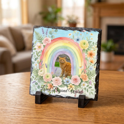 A square slate plaque on a small black stand, featuring a colorful rainbow, a ginger cat, pastel flowers, and the text 'Loved Beyond Words' on a wooden table in a softly blurred living room setting.