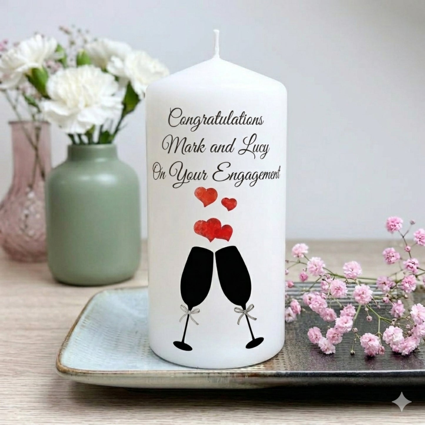 A white cylindrical candle with black script text reading 'Congratulations Mark and Lucy On Your Engagement' and a graphic of two clinking champagne glasses with red hearts, displayed on a tray with flowers in the background.