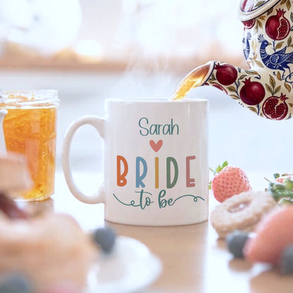 Mug with 'Sarah Bride to Be' text being filled with tea, surrounded by pastries and fruit.