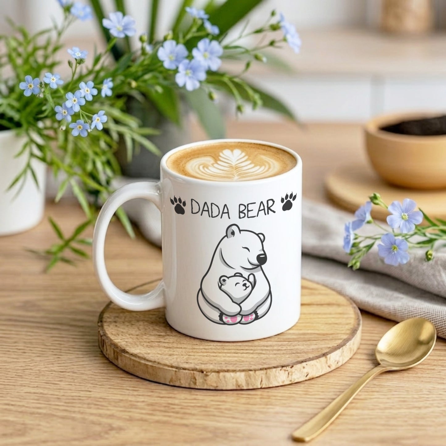 A white ceramic coffee mug with a cute black line art polar bear illustration and the text 'DADA BEAR' on the side, resting on a round wooden coaster. The mug is styled in a cozy cafe-like setting with a latte, blue flowers, a gold spoon, and a neutral fabric background.