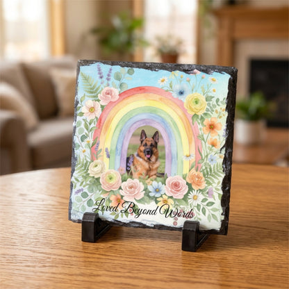 A square slate memorial plaque on a small black display stand, featuring a colorful rainbow, flowers, and a German Shepherd portrait, with the text 'Loved Beyond Words' on a wooden table in a softly blurred living room setting.