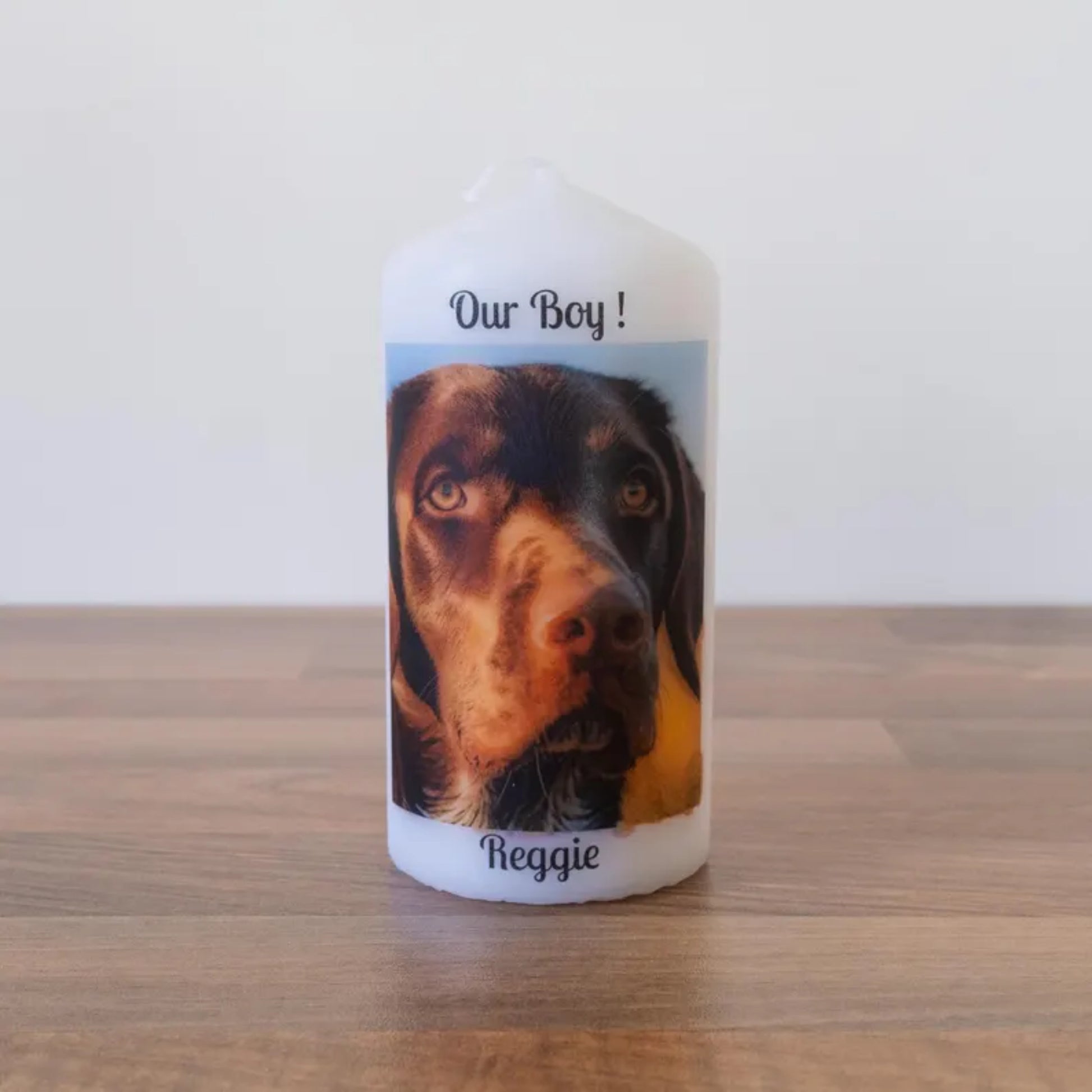 A white pillar candle with a custom printed label featuring a close-up photo of a brown and black dog, with the text 'Our Boy!' at the top and the name 'Reggie' at the bottom, standing on a wooden surface against a plain white wall.