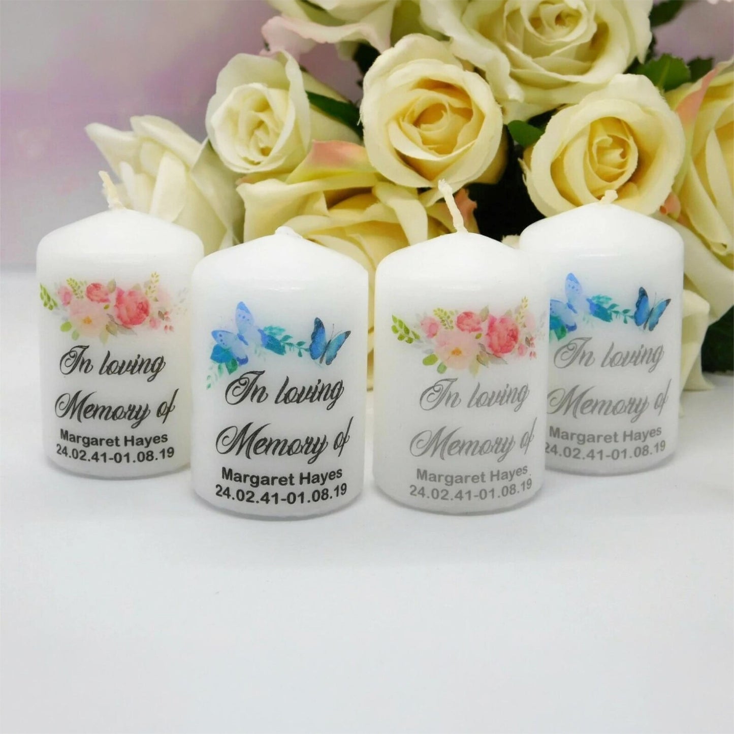 Four white memorial taper candles with floral and butterfly designs, printed with the text 'In loving Memory of Margaret Hayes' and a date of death, arranged in front of a bouquet of cream roses.