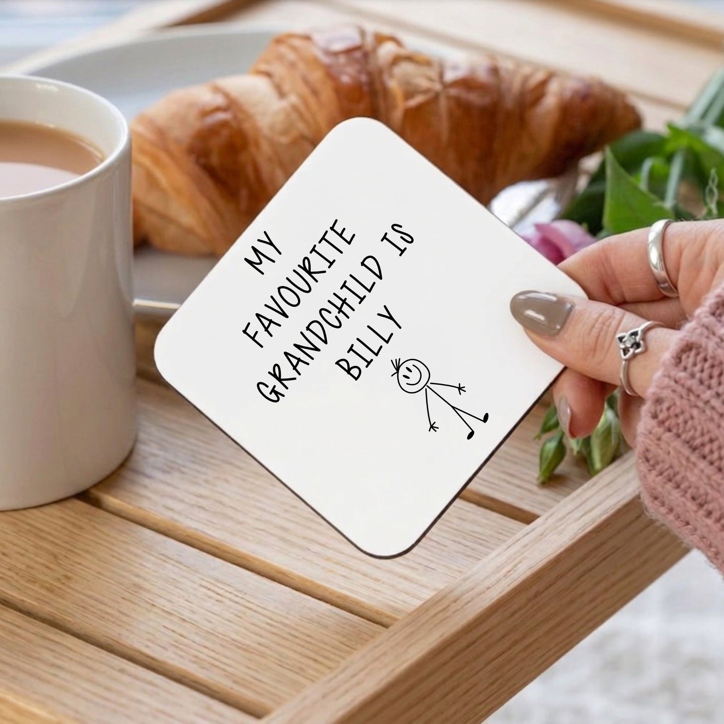 A person holding a white coaster with My favourite grandchild is Billy on it over a breakfast tray with a mug of tea, croissant and rose