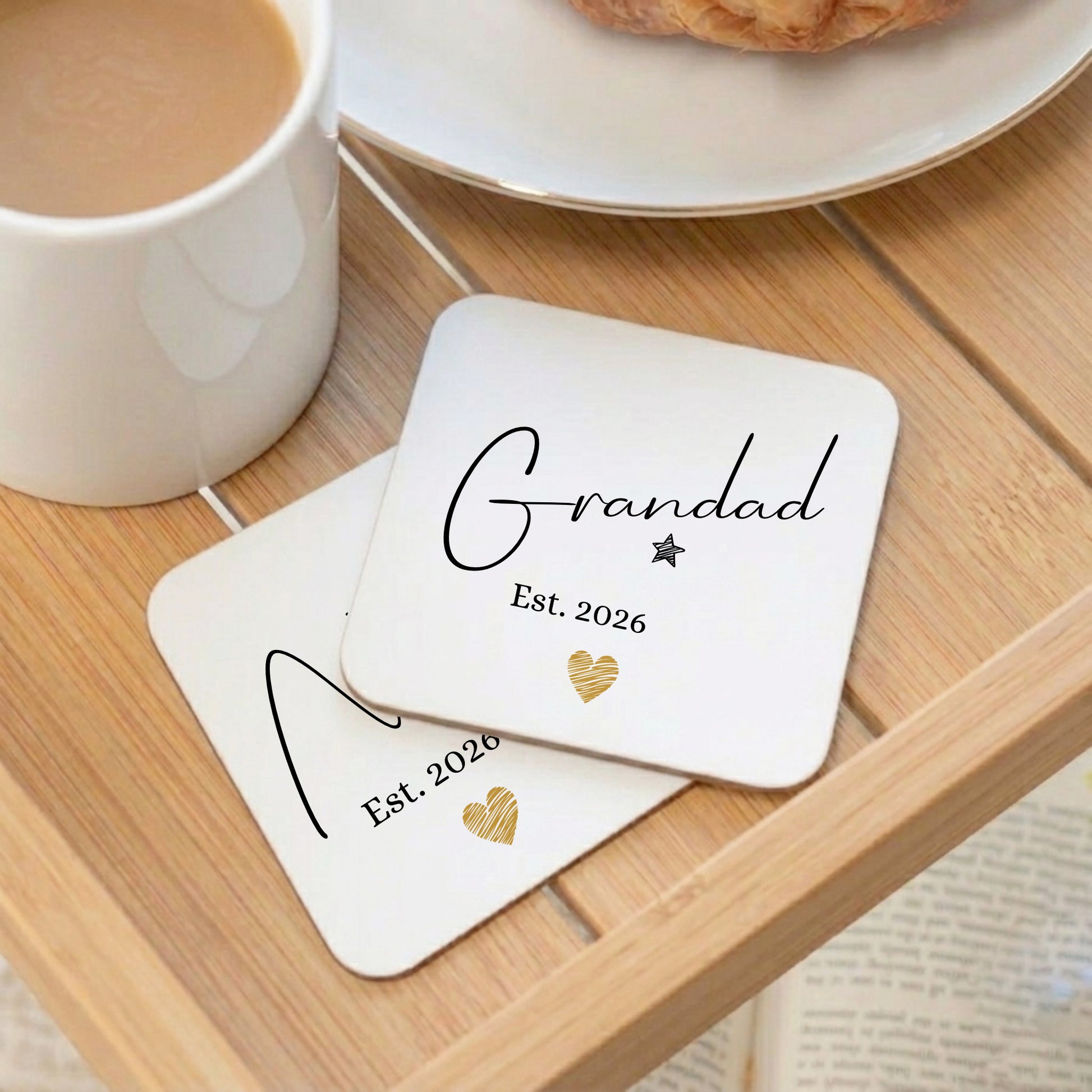 Two coasters with 'Grandad Est. 2026' text on a wooden table with a cup of coffee.