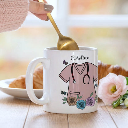 White mug with a design of a nurse's uniform, flowers, and the name 'Caroline'. A hand is holding a gold spoon over the mug.