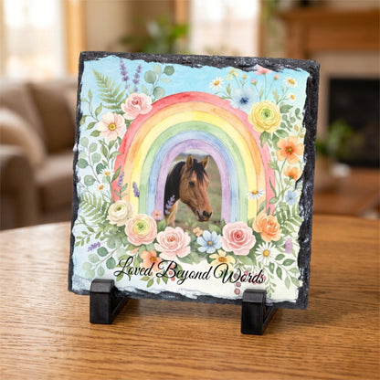 A square slate plaque on a small black display stand, featuring a painted rainbow, flowers, a horse head, and the phrase 'Loved Beyond Words' on a wooden table in a softly blurred indoor setting.