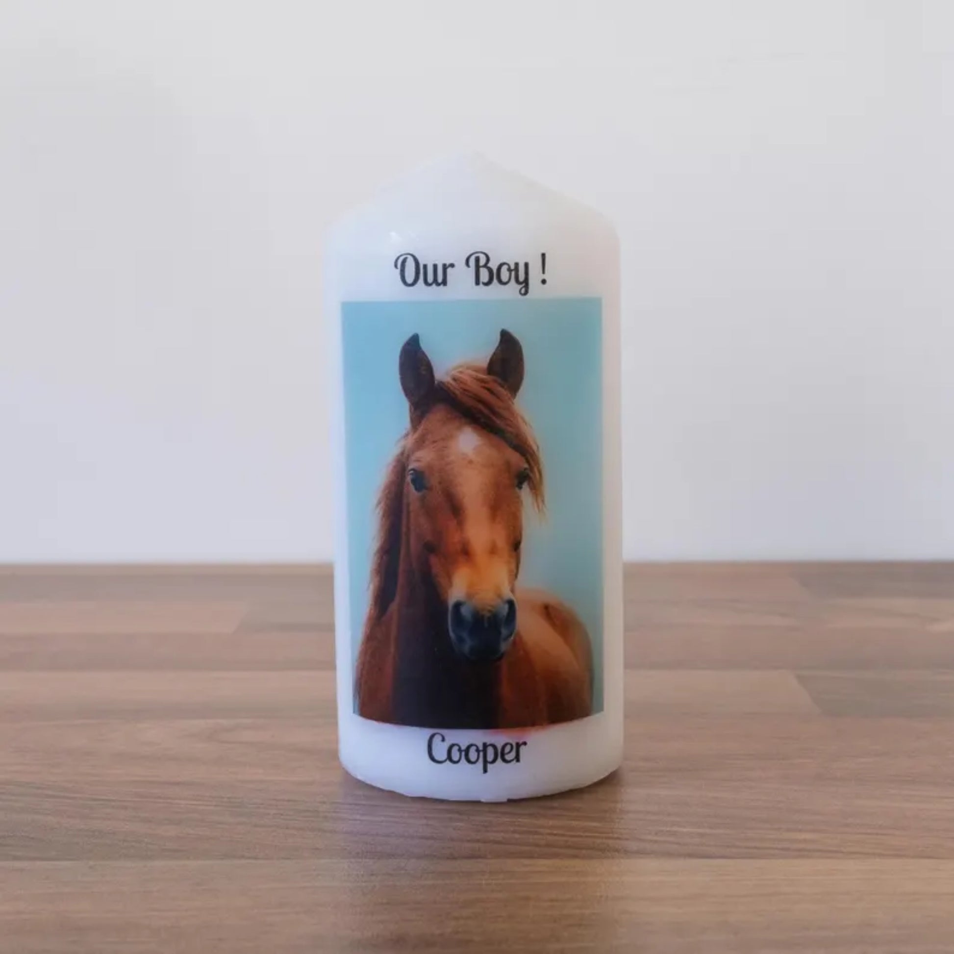 A white custom candle with a printed photo of a brown horse and the text 'Our Boy! Cooper' on the front, standing on a wooden surface against a plain white wall.