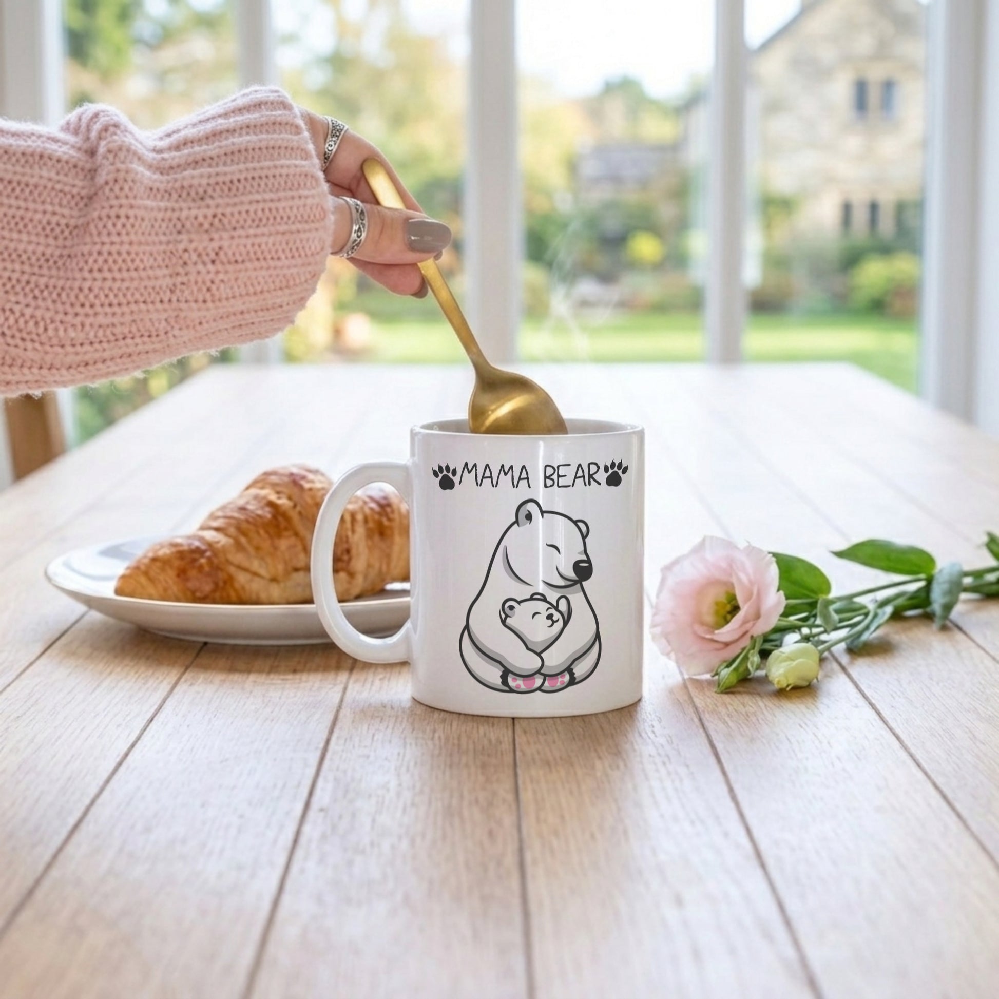 A white ceramic mug with a black line art polar bear illustration and the text 'MAMA BEAR' sits on a wooden table. A hand wearing a pink knit sleeve holds a gold spoon above the mug.