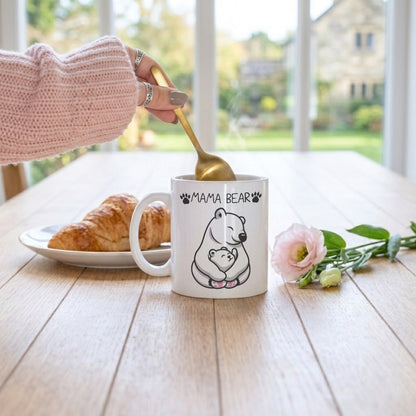 A white ceramic mug with a black line art polar bear illustration and the text 'MAMA BEAR' sits on a wooden table. A hand wearing a pink knit sleeve holds a gold spoon above the mug.