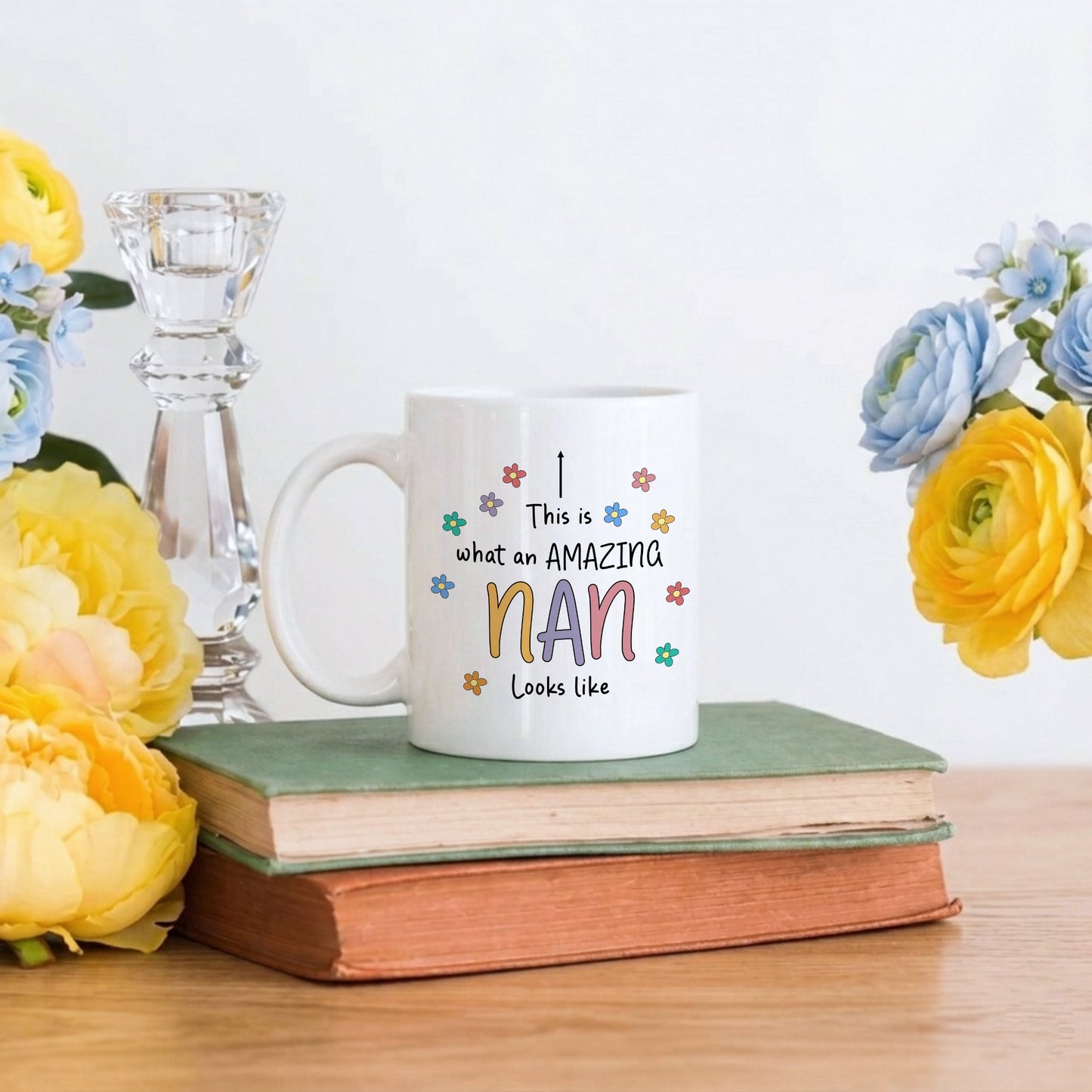 White ceramic mug with the text 'This is what an Amazing Nan looks like' in colourful lettering surrounded by small daisy flowers, displayed on stacked books with yellow and blue flowers
