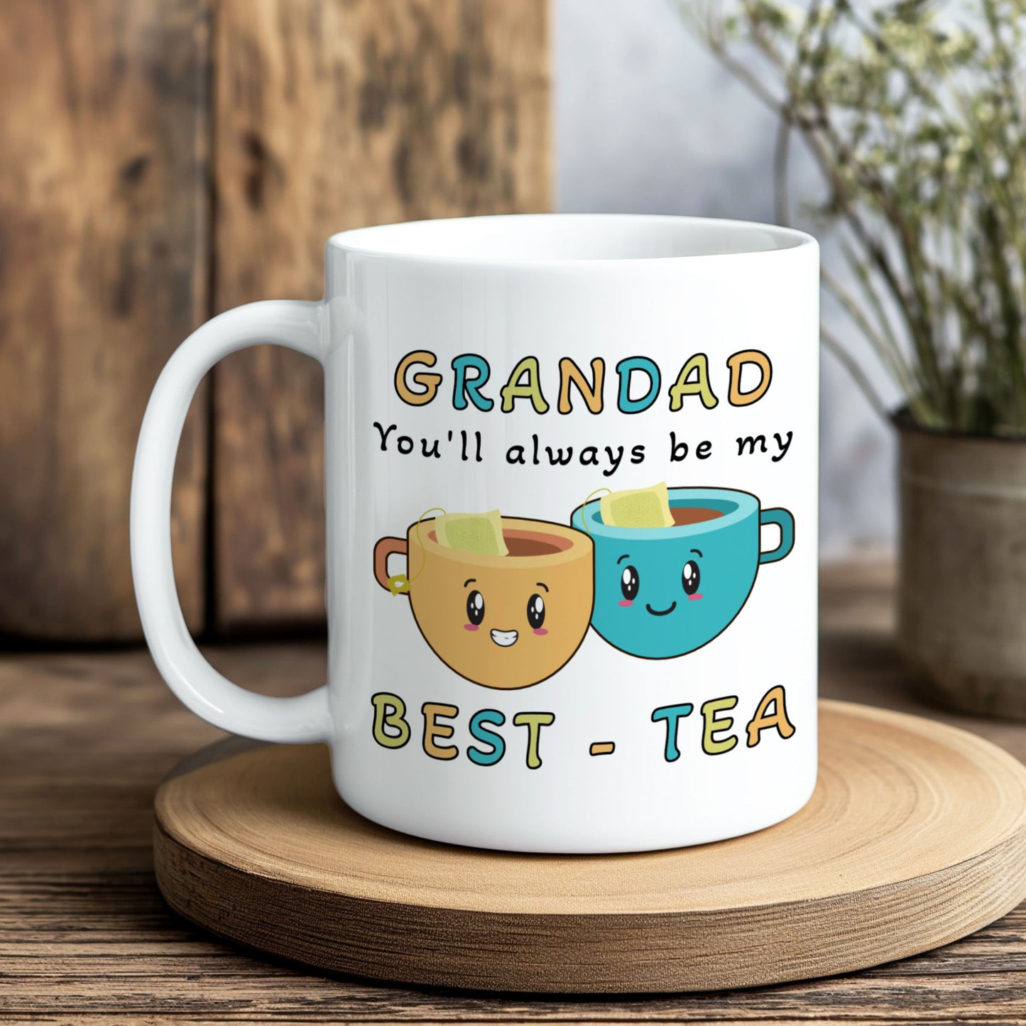 A white ceramic mug with a cartoon graphic of two smiling tea cups and the text 'GRANDAD You'll always be my BEST - TEA', displayed on a wooden coaster with a rustic wooden background.