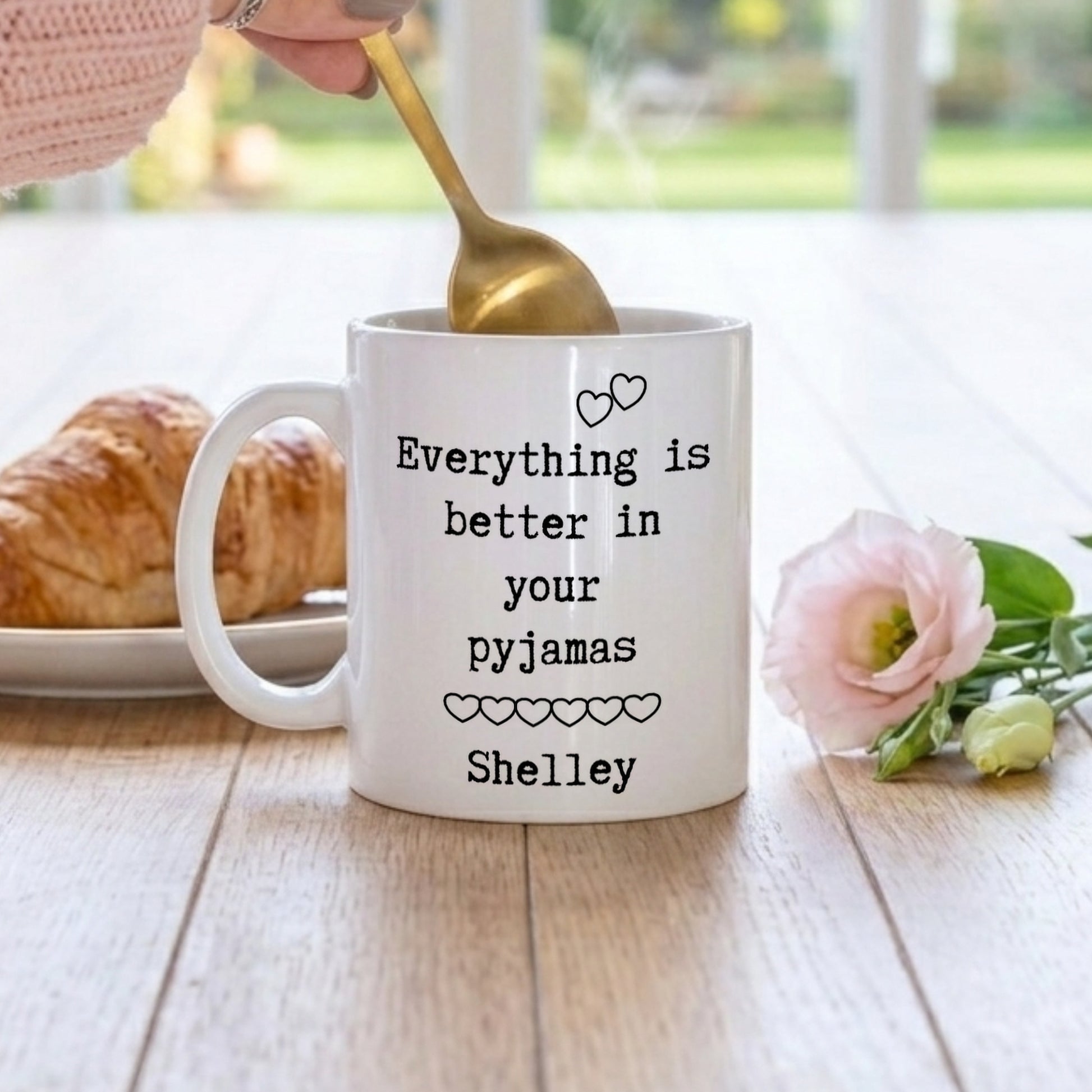 A white ceramic mug with black text reading 'Everything is better in your pyjamas Shelley' sits on a wooden table. A hand holding a gold spoon stirs the mug, with a croissant on the left and a pink flower on the right, against a softly blurred background.