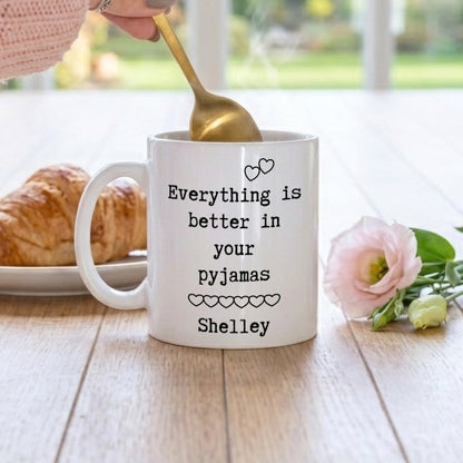 A white ceramic mug with black text reading 'Everything is better in your pyjamas Shelley' sits on a wooden table. A hand holding a gold spoon stirs the mug, with a croissant on the left and a pink flower on the right, against a softly blurred background.