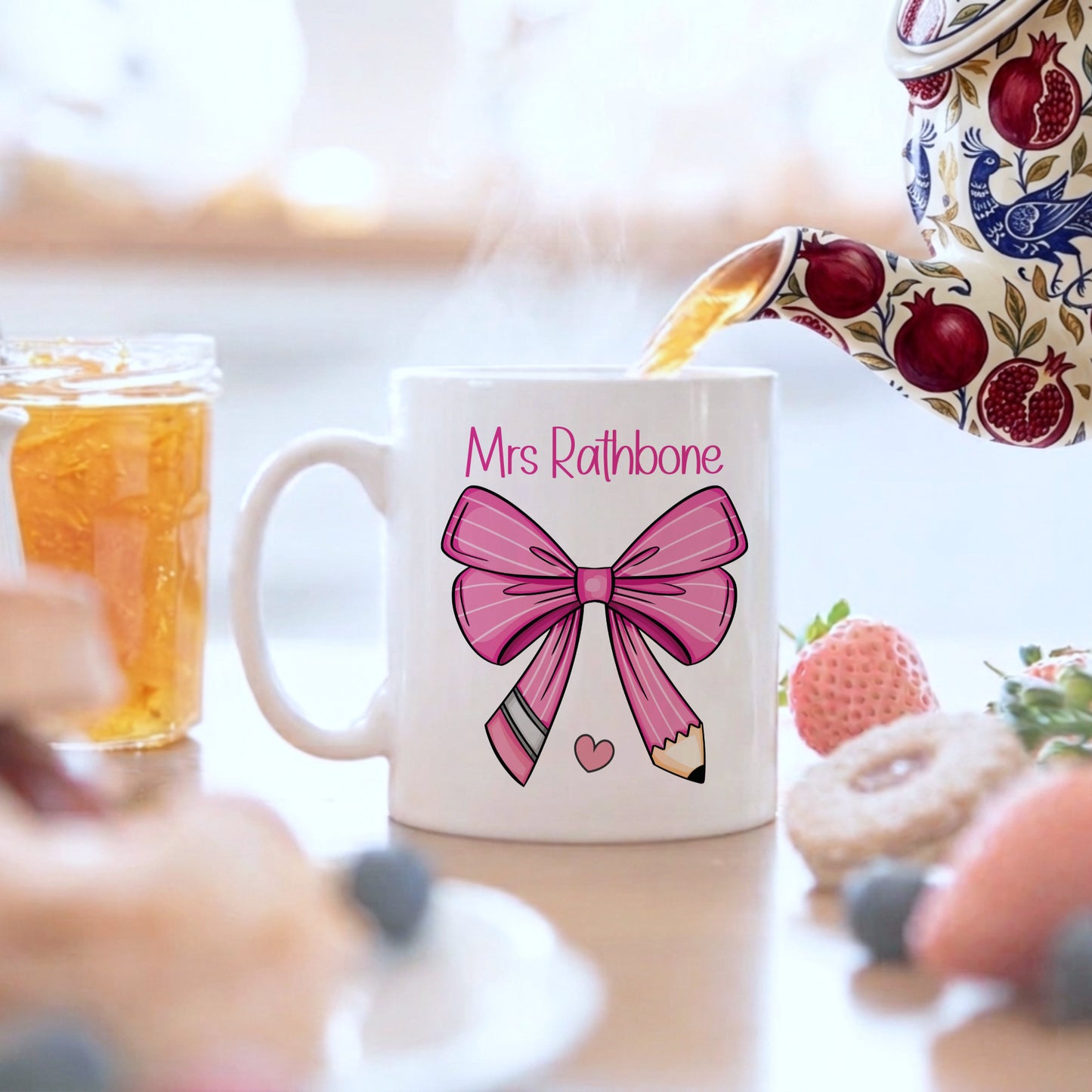 Personalised teacher mug with a pink bow and pencil design, customised with 'Mrs Rathbone', being filled from a floral teapot alongside iced tea and fresh fruit