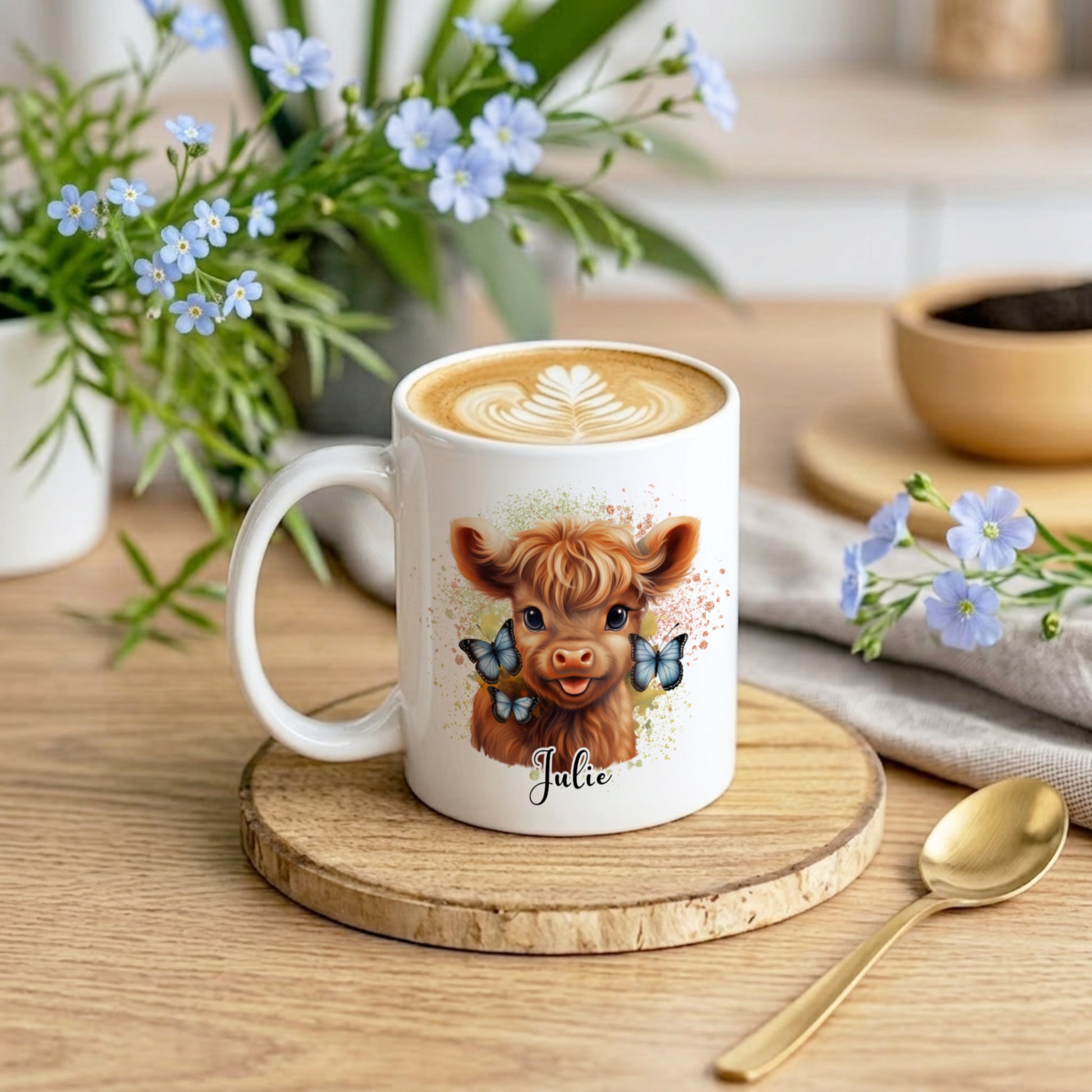 White mug with a cow illustration and 'Julie' text on a wooden coaster with flowers and a spoon.