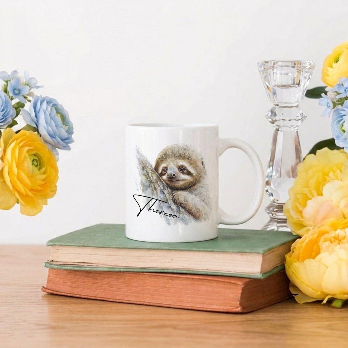 Mug with a sloth design on a stack of books with flowers and a candle in the background