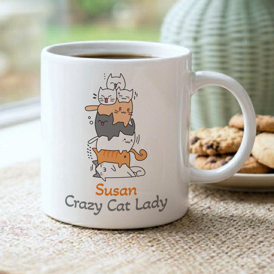 White ceramic mug with a stack of cartoon cats in different colours. The mug has a curved handle and the text "Susan Crazy Cat Lady" printed on the front. The mug is on a neutral-coloured surface.