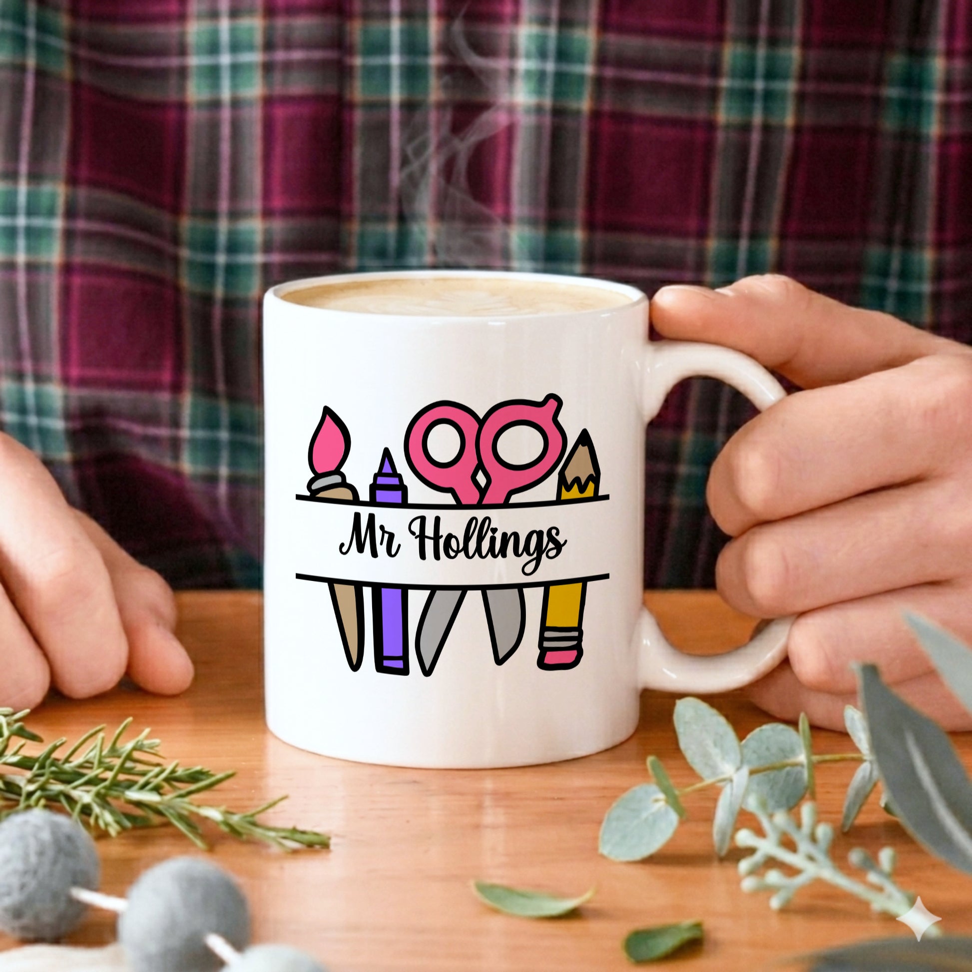 Person holding a white mug with personalized design and text, surrounded by decorative elements on a wooden surface.