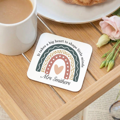 Coaster with rainbow design and text on a wooden table with a mug and flowers.