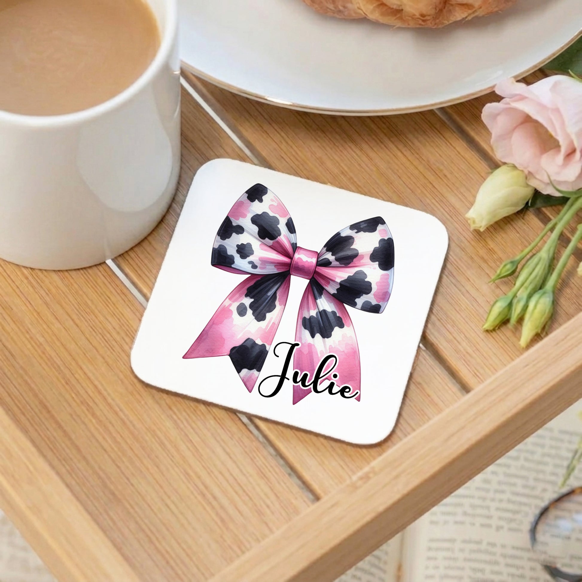 Personalised 'Julie' coaster with a pink and black cow print bow design, displayed on a wooden tray alongside a white mug of coffee and pink flowers