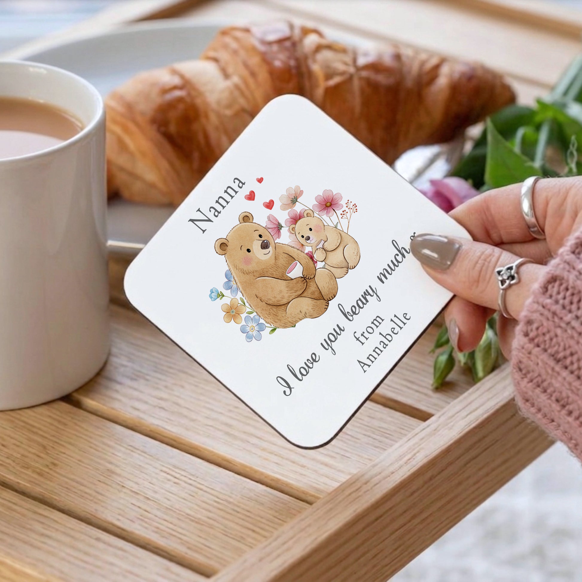 Coaster with teddy bears and personalized message on a wooden tray with coffee and croissant.