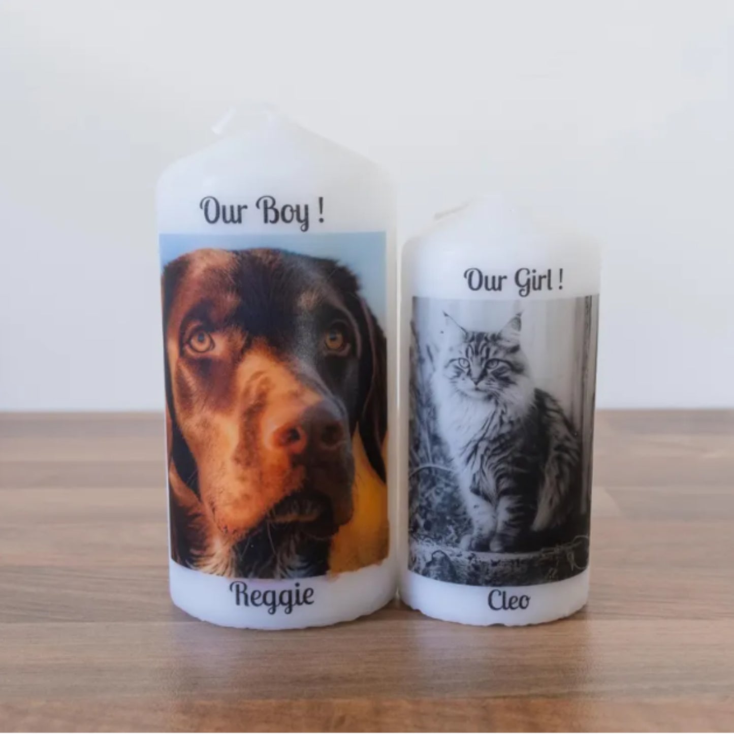 Two personalised white taper candles standing on a wooden surface against a plain light background. The left candle is labelled 'Our Boy!' with a photo of a brown and black dog named Reggie, and the right candle is labelled 'Our Girl!' with a black-and-white photo of a cat named Cleo.