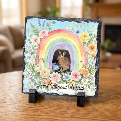 A square decorative slate plaque on a small black display stand, featuring a colorful rainbow, flowers, a rabbit, and the phrase 'Loved Beyond Words' on a wooden table in a softly blurred living room setting.