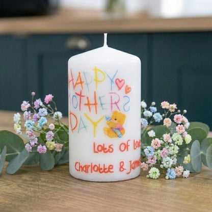 White candle with colorful text and a teddy bear illustration, surrounded by flowers on a wooden surface.