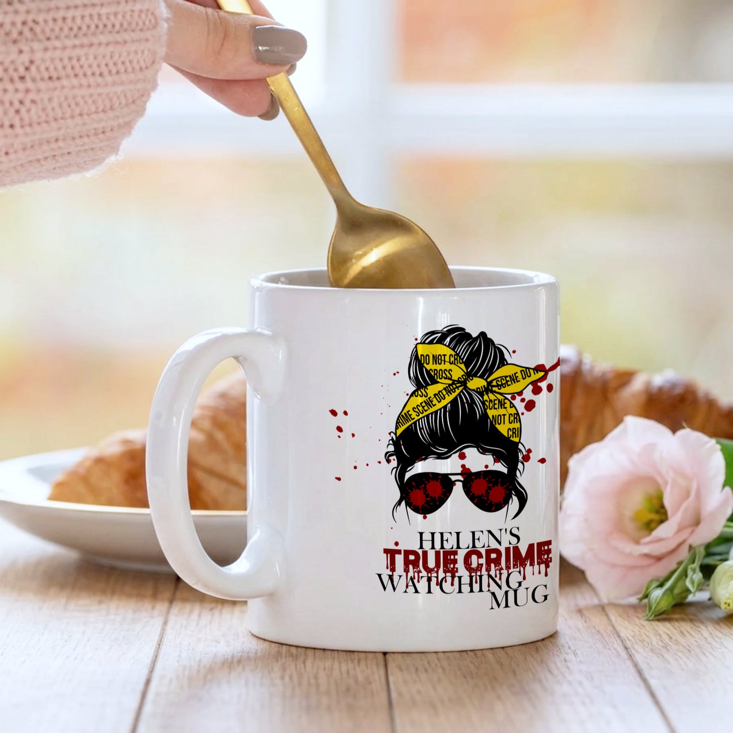 White mug with a design and text, being held with a spoon, on a table with a blurred background.