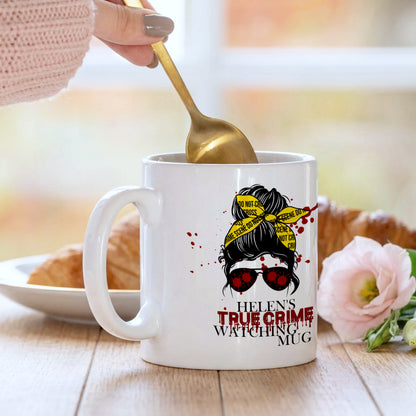 White mug with a design and text, being held with a spoon, on a table with a blurred background.