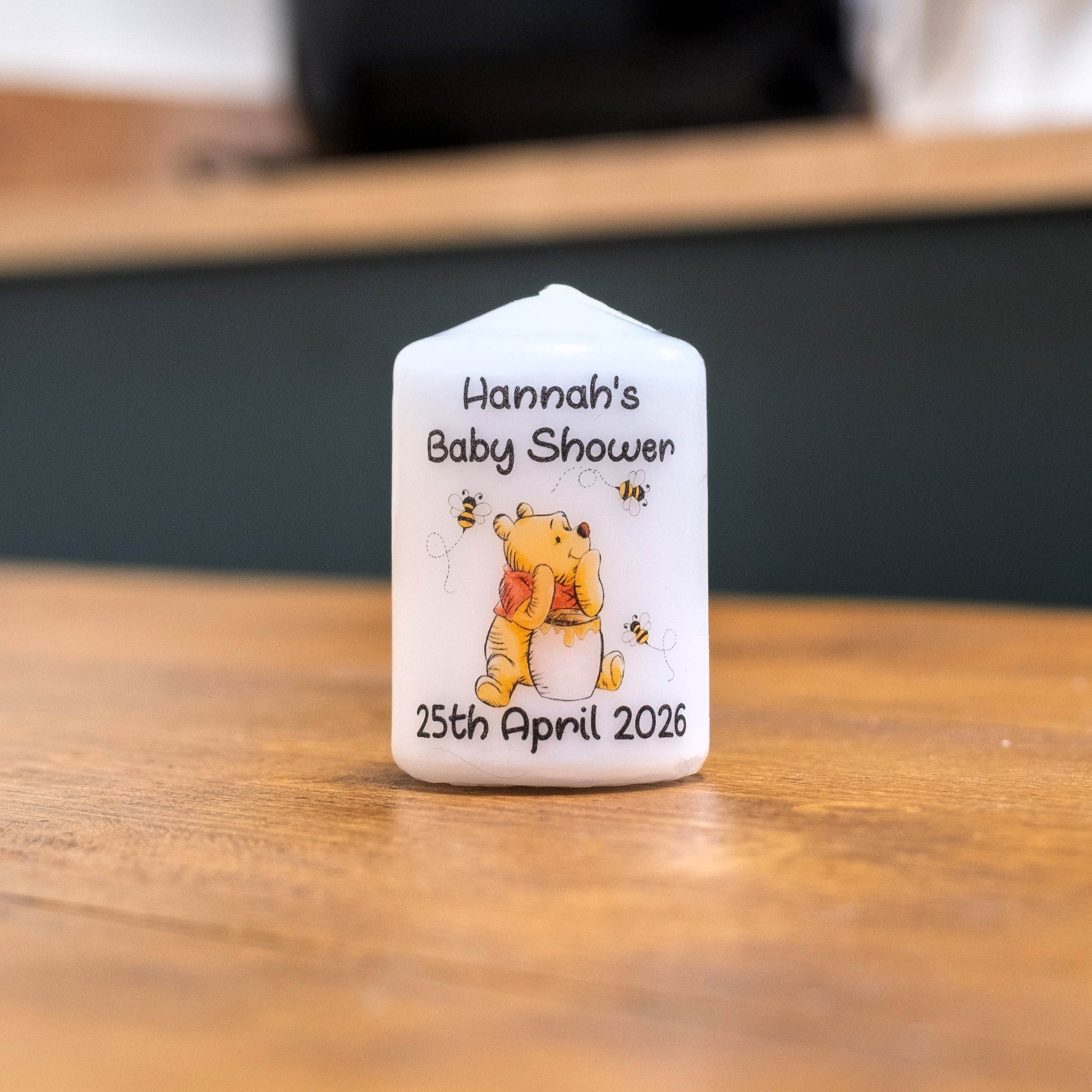 A custom white candle featuring Winnie the Pooh printed on the front, personalised with the text 'Hannah's Baby Shower' and the date '25th April 2026'. The candle is shown standing on a wooden tabletop with a blurred background.