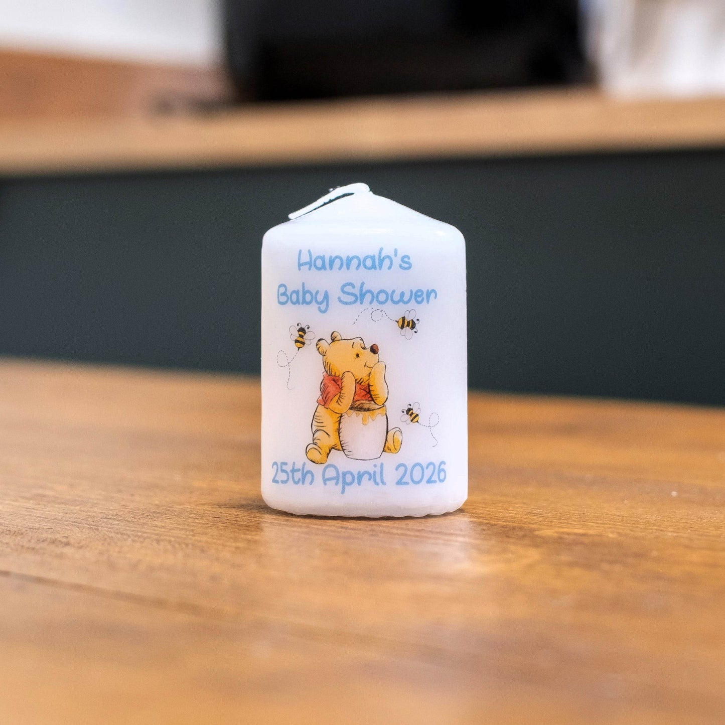 A white personalised baby shower candle featuring Winnie the Pooh and bees, with the text 'Hannah's Baby Shower' and the date '25th April 2026', sitting on a wooden table.