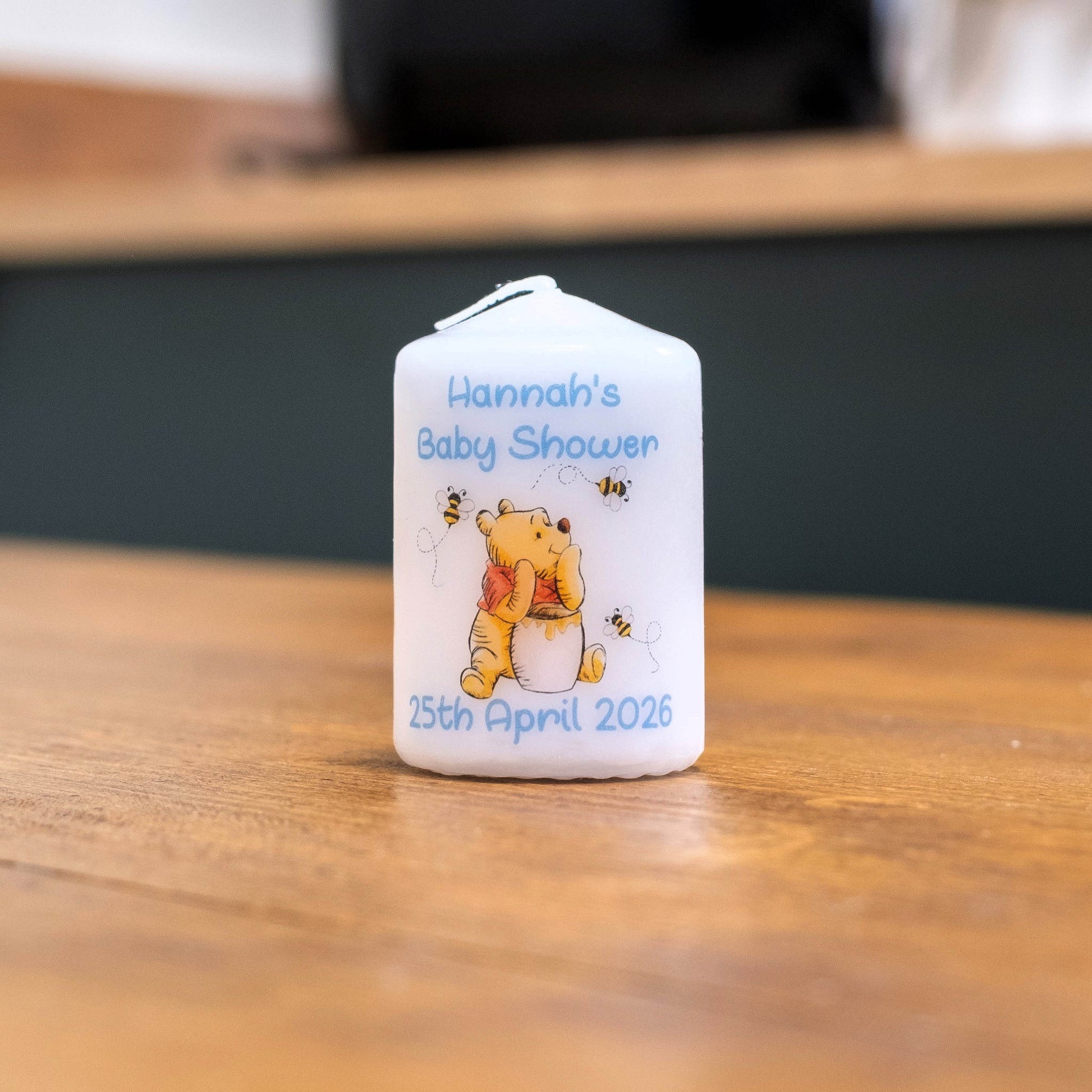 A white personalised baby shower candle featuring Winnie the Pooh and bees, with the text 'Hannah's Baby Shower' and the date '25th April 2026', sitting on a wooden table.