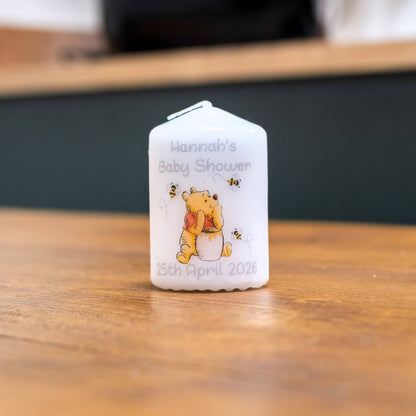 A small white baby shower candle favour on a wooden table, printed with the text 'Hannah's Baby Shower 25th April 2026' and an illustration of Winnie the Pooh holding a honey pot, with bees around it.