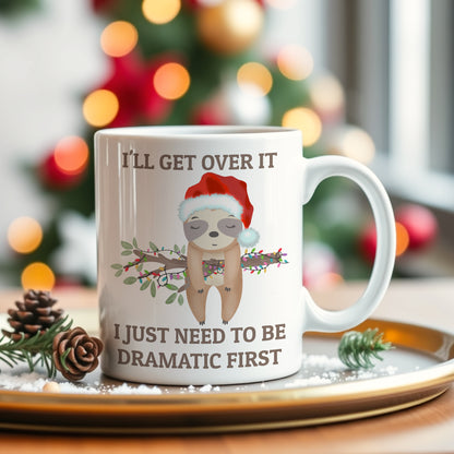 White mug with a sloth wearing a Santa hat and Christmas-themed text, placed on a decorative tray with a blurred Christmas tree in the background.