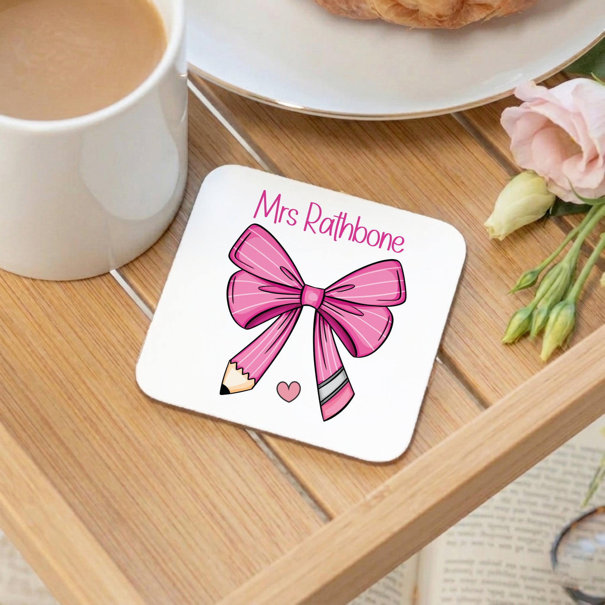 Personalised 'Mrs Rathbone' teacher coaster with a pink bow and pencil design, displayed on a wooden tray alongside a white mug and pink flowers