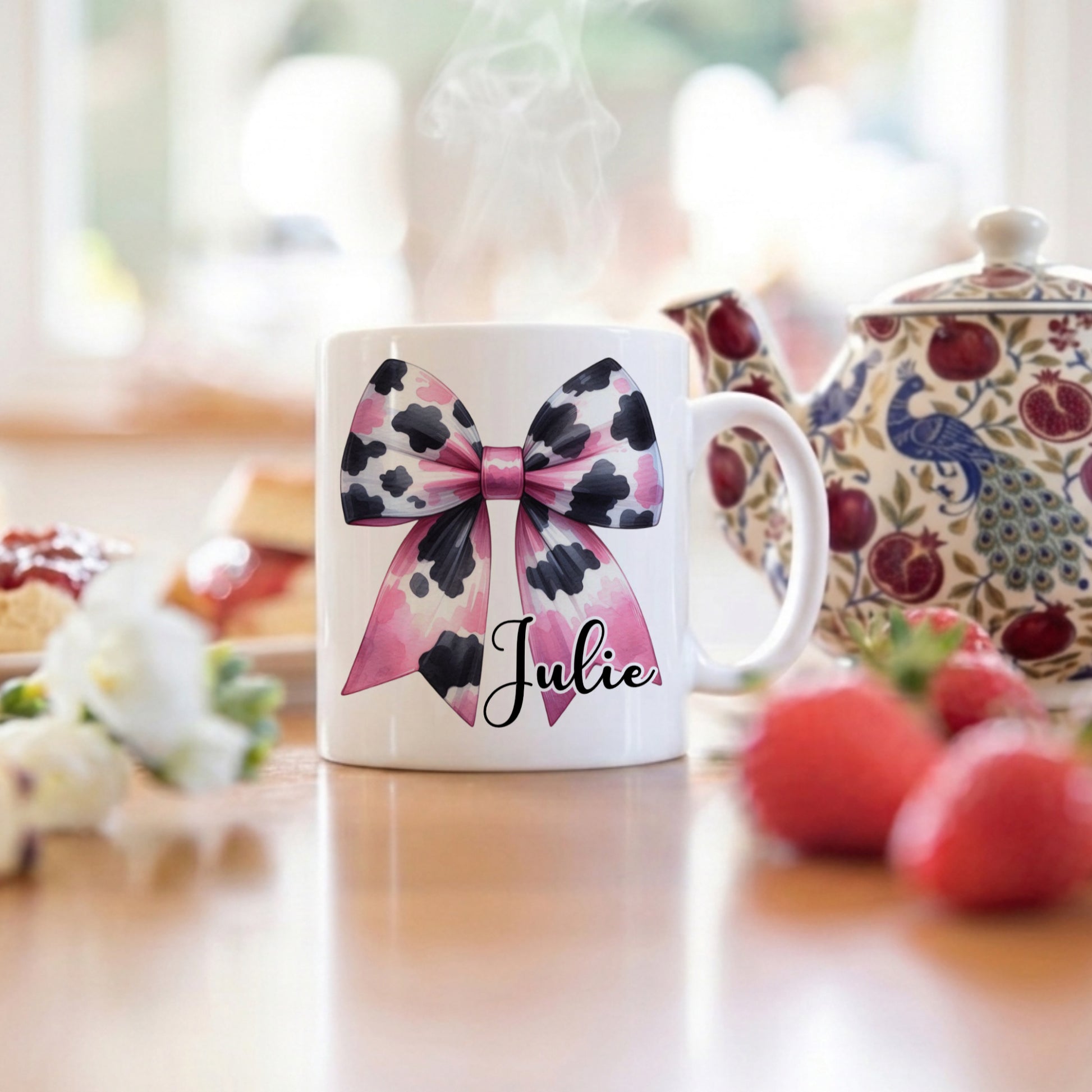 Personalised 'Julie' mug with a pink and black cow print bow design, displayed on a table alongside a floral teapot and fresh strawberries